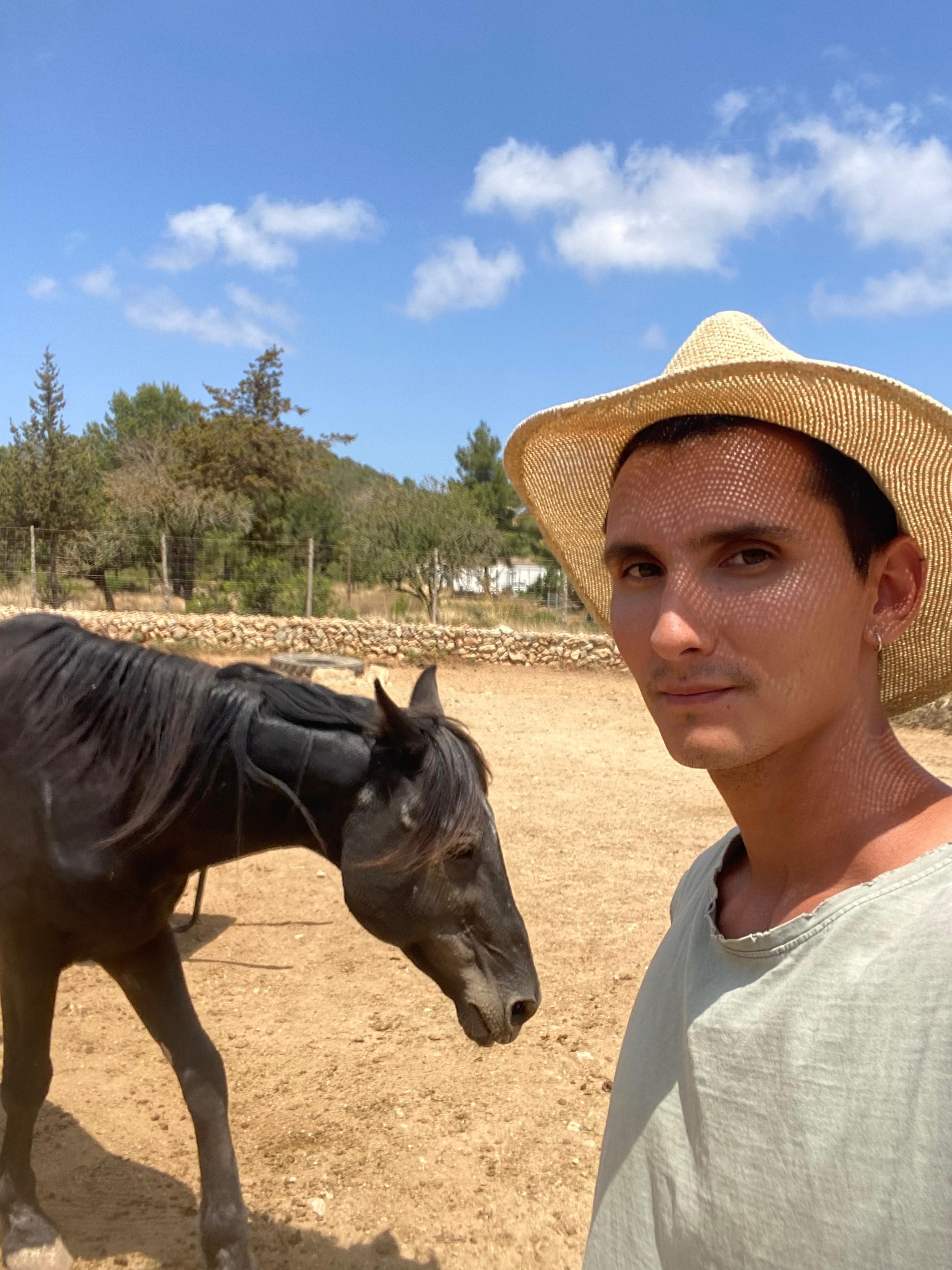 Izhy con un cappello di paglia accanto a Padre, un cavallo nero, sulla terra secca vicino a un muro a secco, dietro pini e cielo azzurro.