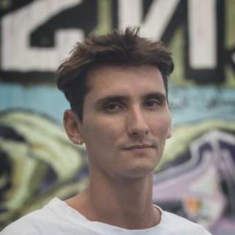 Izhy, short dark hair and a white T-shirt, in front of a large graffiti wall.