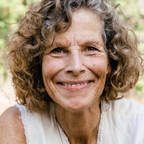 Portrait of Lisbeth La Fors smiling warmly, curly hair, a white sleeveless top, in green sunlit leaves.
