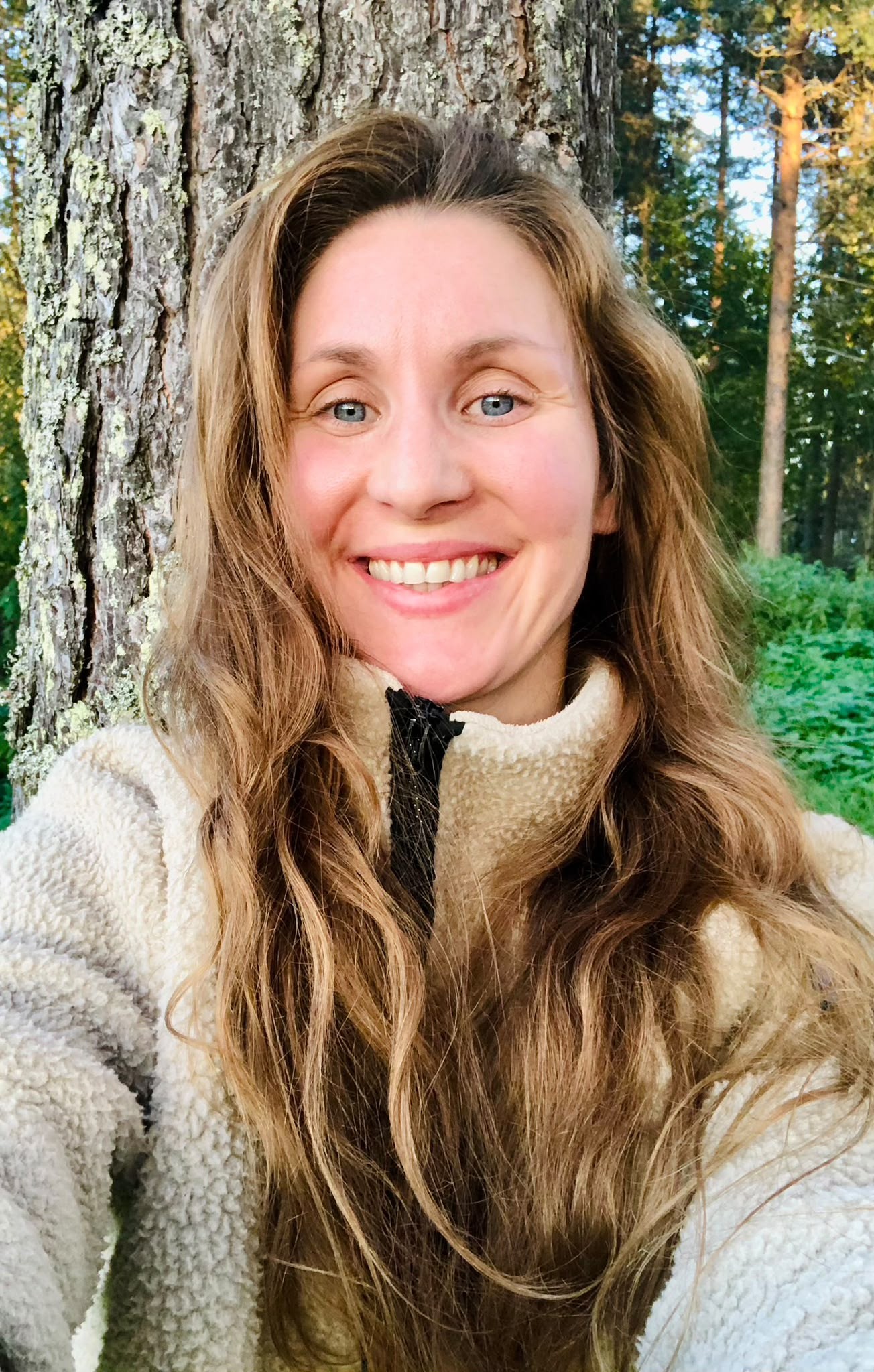 Martina Strand smiling, long wavy light-brown hair, in a cream fleece, leaning against a pine trunk in a sunlit forest.