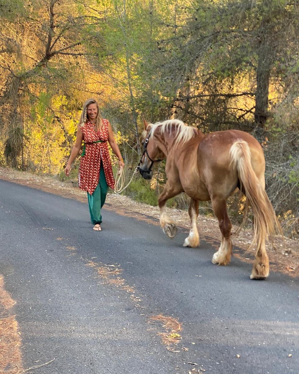 Marzena walks down a quiet road in evening light beside Madre, a chestnut mare with a blond mane, the lead rope loose in her hand, pines and golden trees behind.
