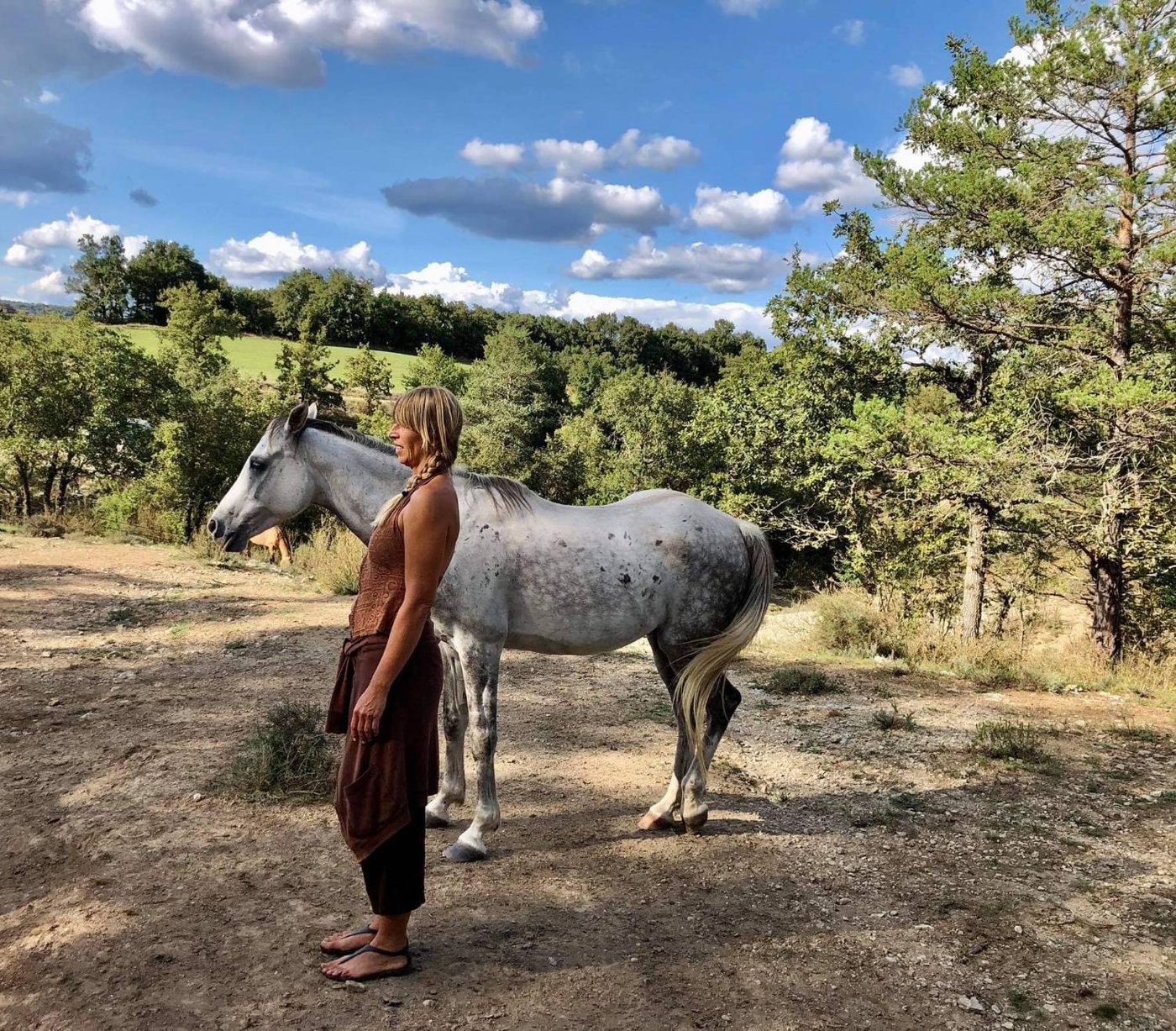 Marzena stands quietly beside a dapple-grey horse on a dry, open track, with pine trees and a green hill behind them.