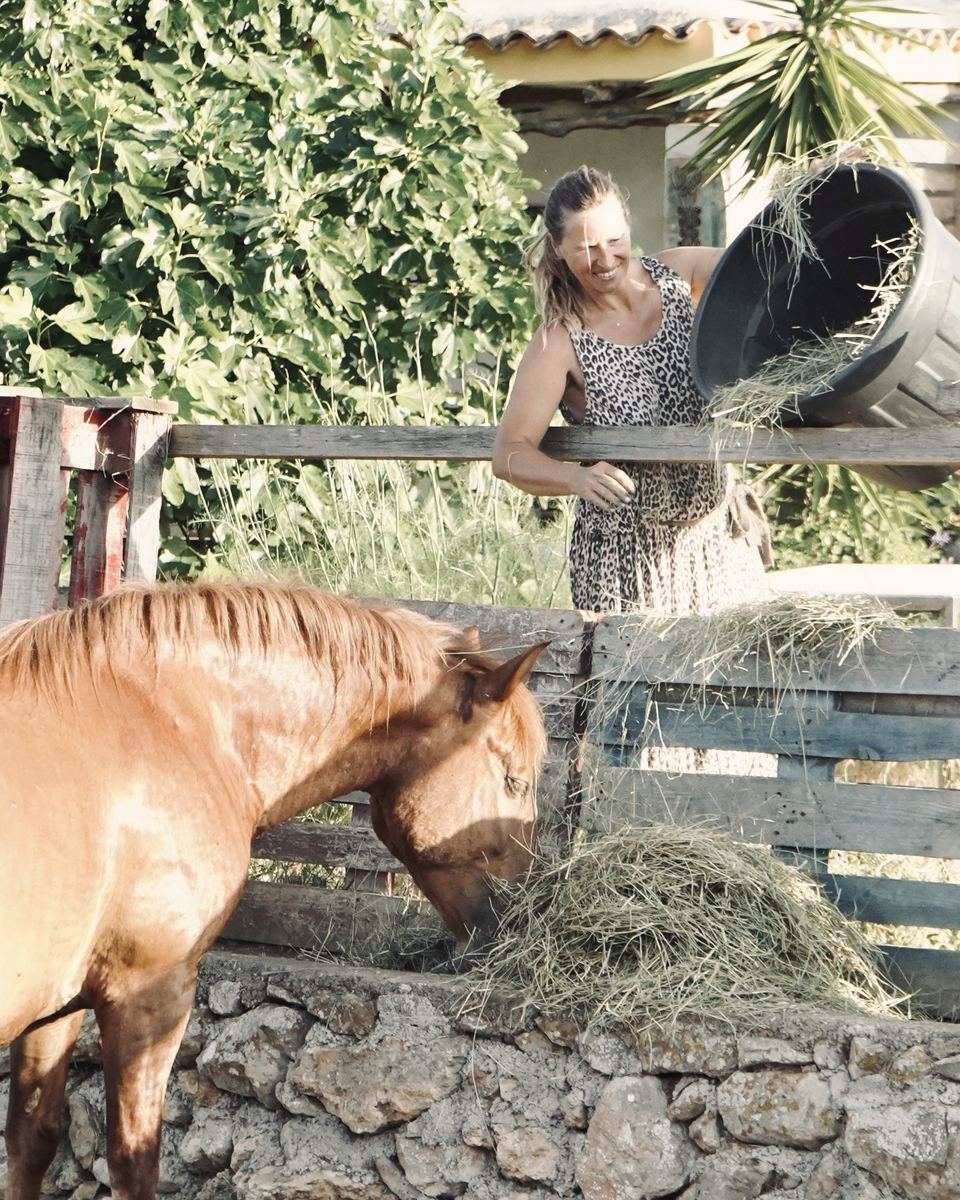 Marzena tips a tub of hay over a wooden fence, laughing, while Madre reaches down to the pile below.