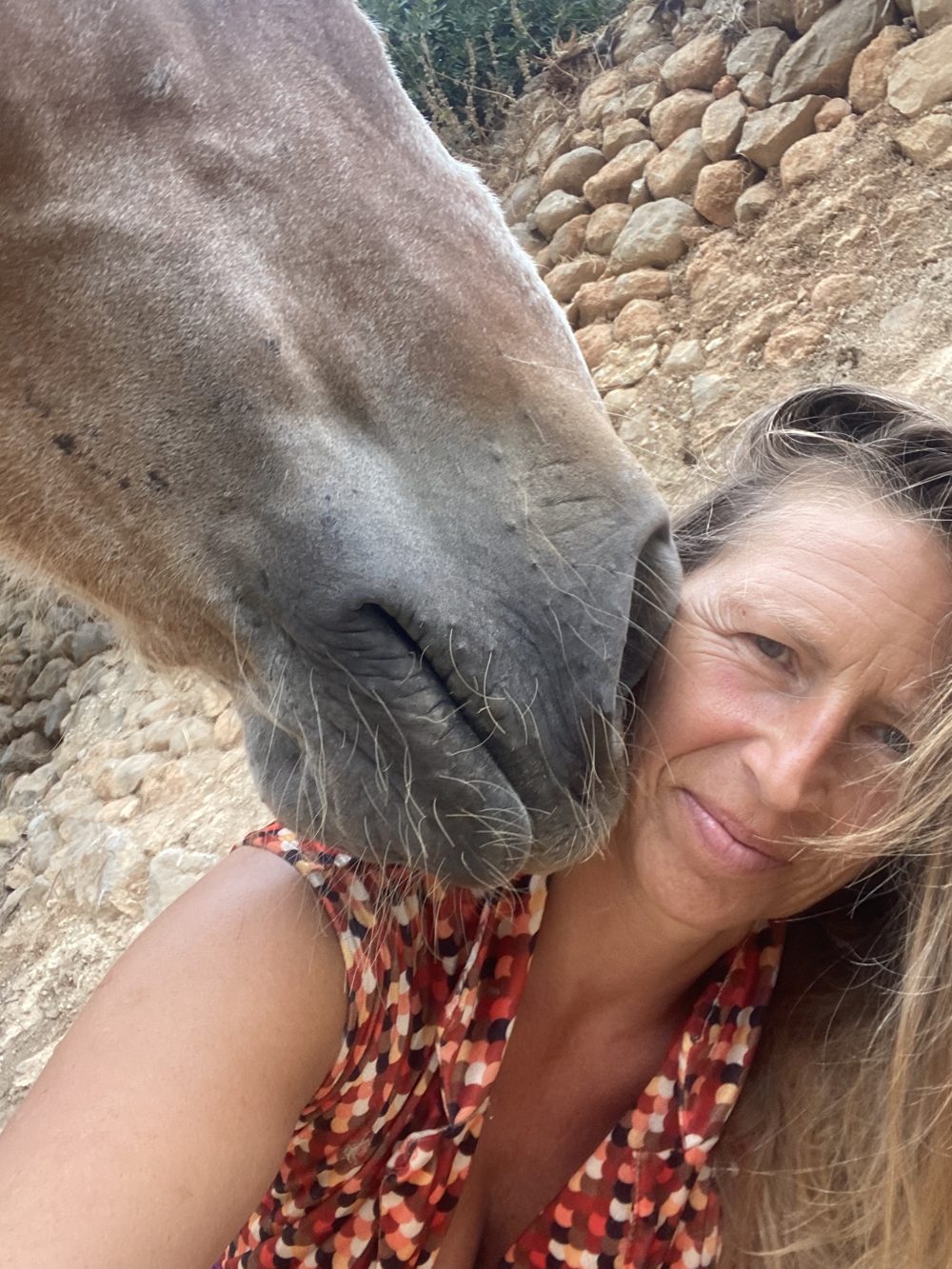Marzena, founder of Horse Presence, smiling softly as a horse rests its muzzle against her cheek, a dry-stone wall behind, at Our Home Can Tigres in Ibiza.