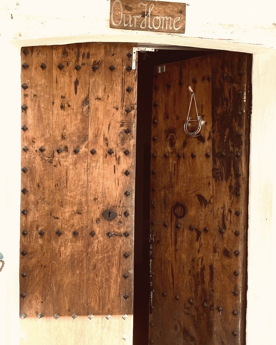 An old studded wooden door standing half open in a whitewashed wall, a horseshoe hanging on a string, and above it a hand-carved sign reading Our Home.
