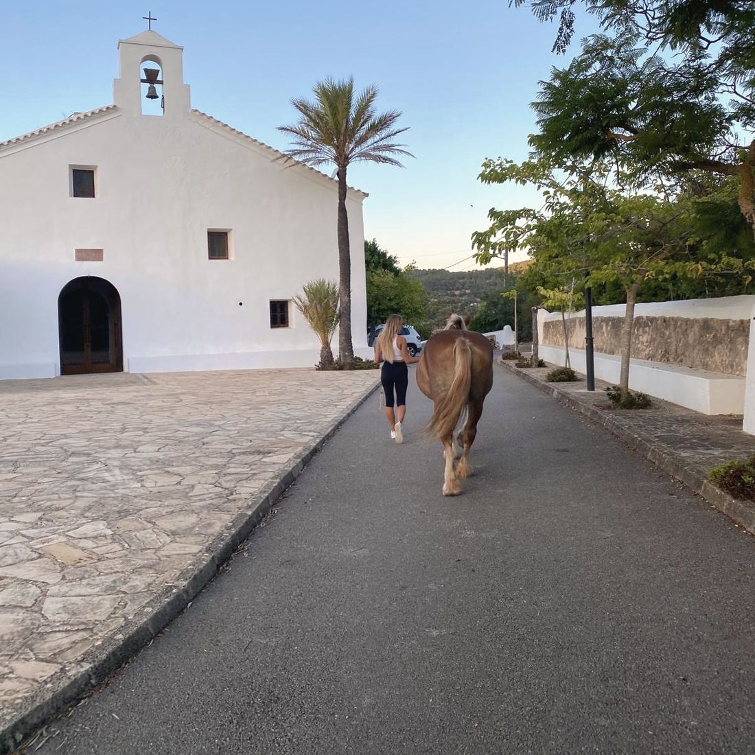 Seen from behind, a woman walks up a village lane beside Madre, past a whitewashed church with a bell tower.