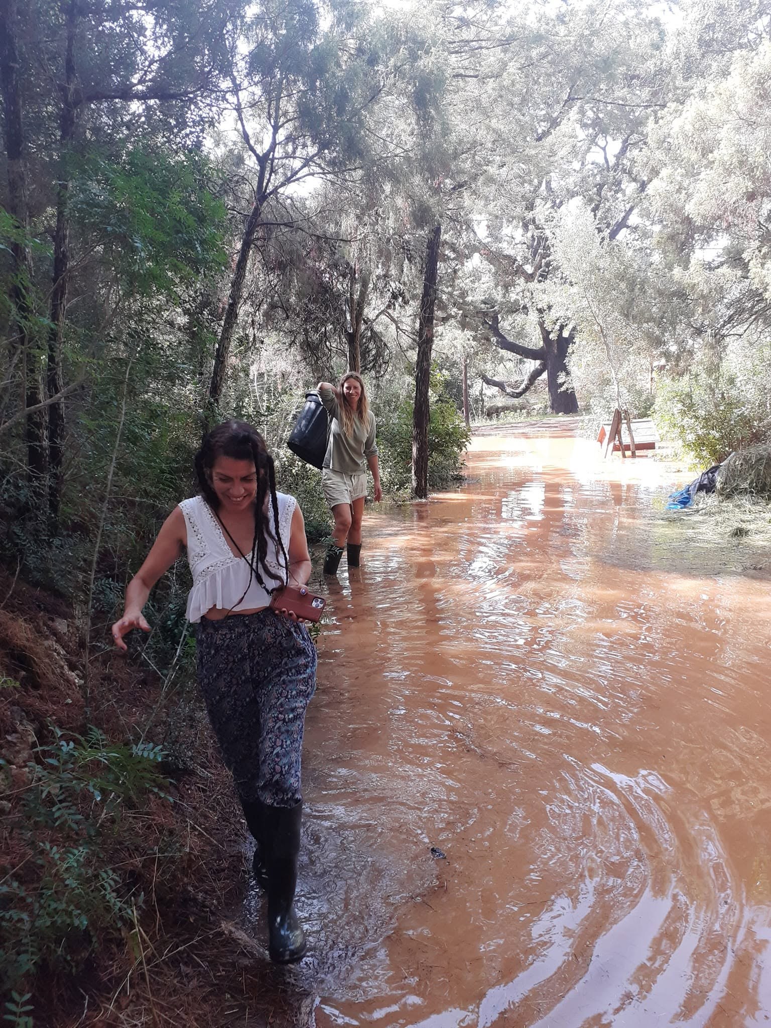 Two women in rubber boots laugh as they wade along a path flooded by the torrent, red-brown water flowing between pines and an old carob tree.